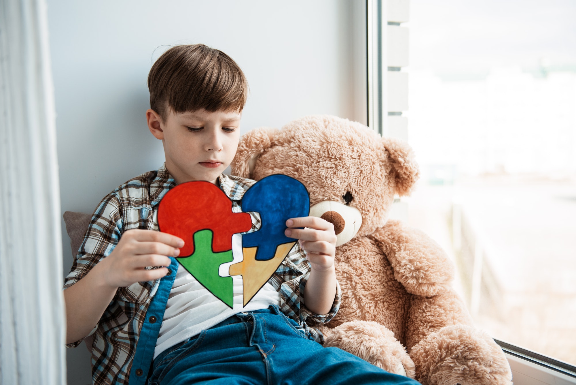 Lonely little boy with teddy bear sitting at home. A Autistic child holds a heart World Autism day