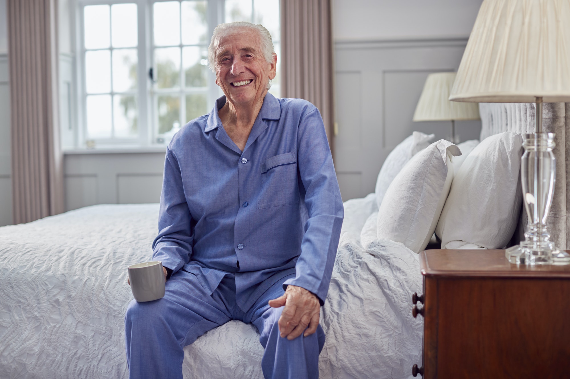 Senior Man At Home Wearing Pyjamas Sitting On Bed Having Morning Cup Of Tea